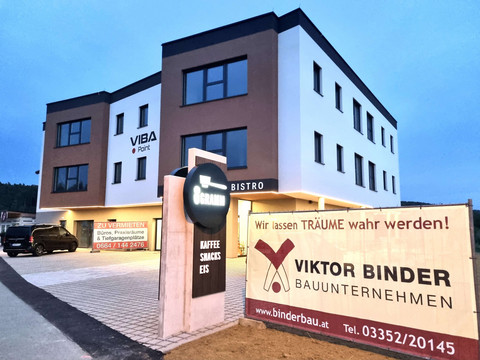 VIBA Point Gewerbezentrum in Kemeten in der Abenddämmerung - modernes dreigeschossiges Gebäude mit Cafe 8 GRAMM im Erdgeschoss und Binder Bau Bauunternehmen Banner mit dem Slogan 'Wir lassen TRÄUME wahr werden!