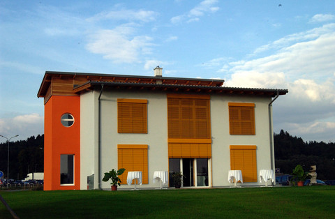 Firmenhauptsitz Kemeten im Jahr 2005, ein modernes Passiv-Musterhaus mit weißer Fassade, orangefarbenem Akzent und gelben Fensterläden. Das Gebäude steht in einem neu erschlossenen Gewerbegebiet und ist von einer gepflegten Grünfläche umgeben. 